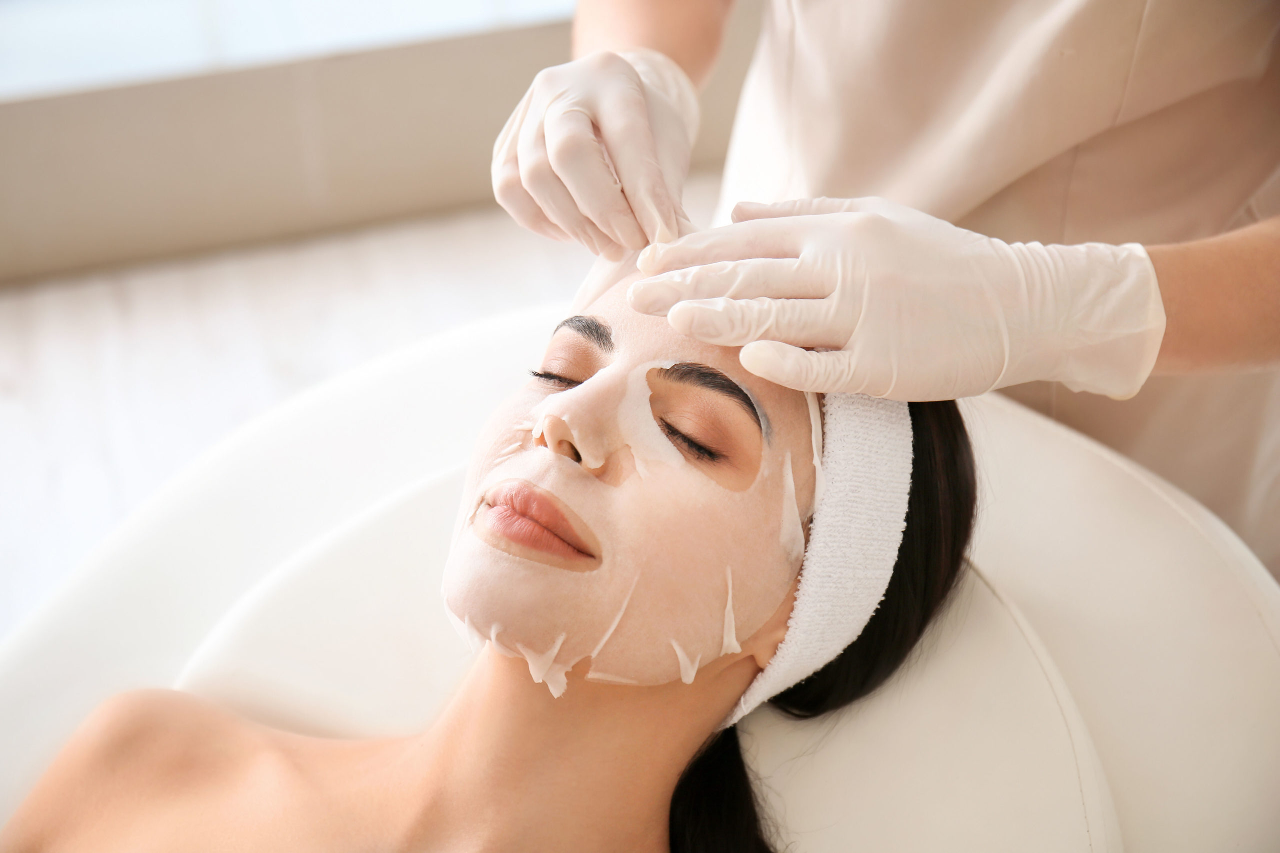 Cosmetologist applying sheet mask on woman's face in beauty salon